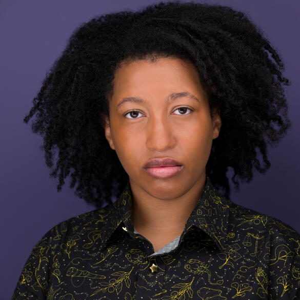 Rhys Tirado A headshot of a person with shoulder-length curly dark hair, brown skin, and a patterned shirt. They are standing in front of a purple backdrop.