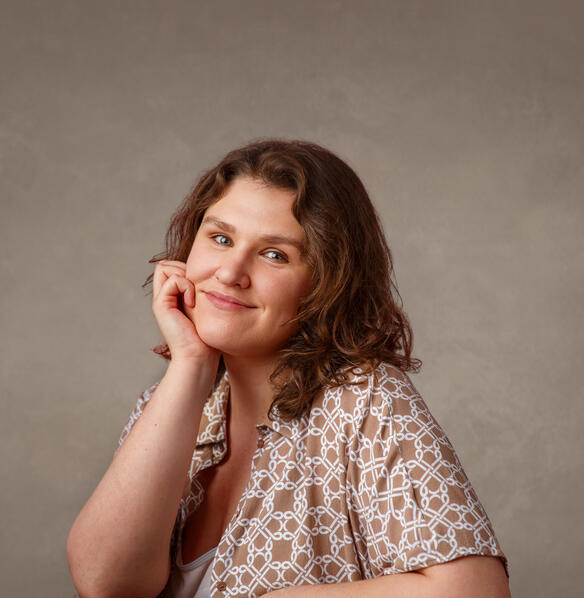 Amelia Stephenson A photo of a person with light skin and brown shoulder-length hair. She is wearing a patterned light brown and white shirt. She is smiling.