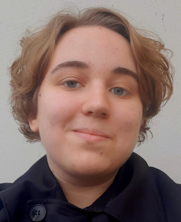 Lumi Oakes A headshot of a person with short blonde hair and light skin. They are wearing a black collared jacket and is standing in front of a white backdrop.