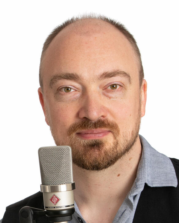 David Ault A headshot of a person with light skin and a short brown beard and brown hair. He is standing in front of a white background and behind a microphone. He is smiling.